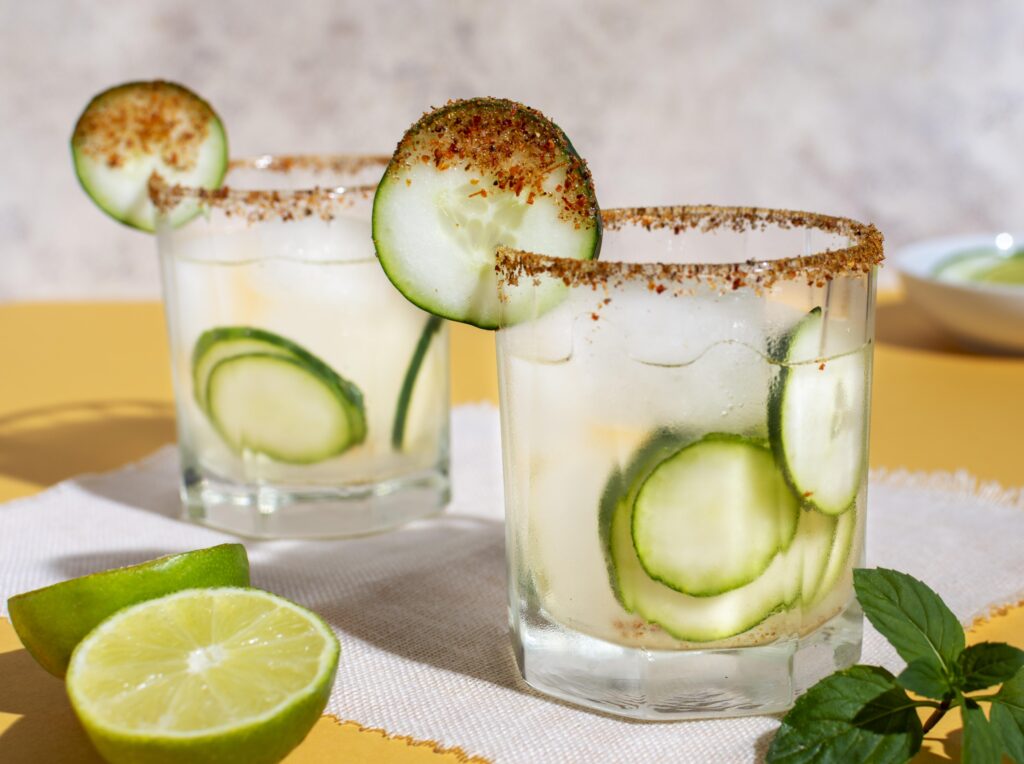 Two glasses of cucumber skinny margarita with a chili-salt rim, garnished with cucumber slices, lime, and fresh mint on a bright background.