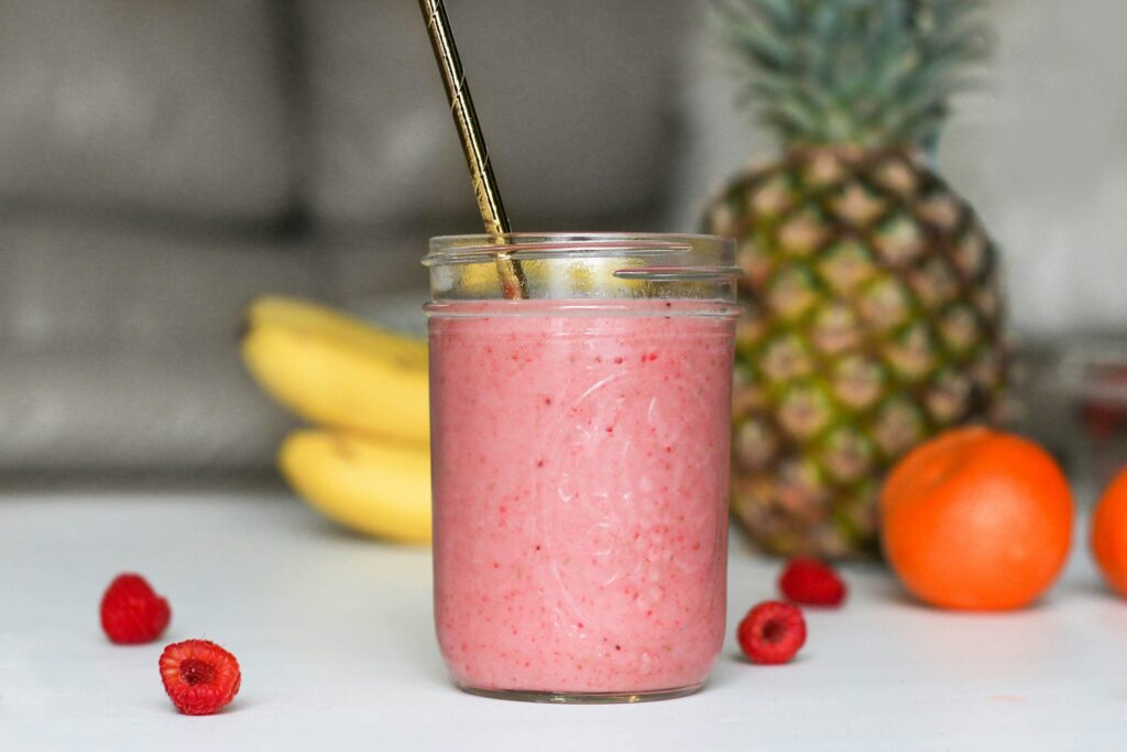 Hailey Bieber Smoothie Recipe featuring a pink smoothie in a jar with a gold straw, surrounded by fresh ingredients like bananas, raspberries, pineapple, and oranges.