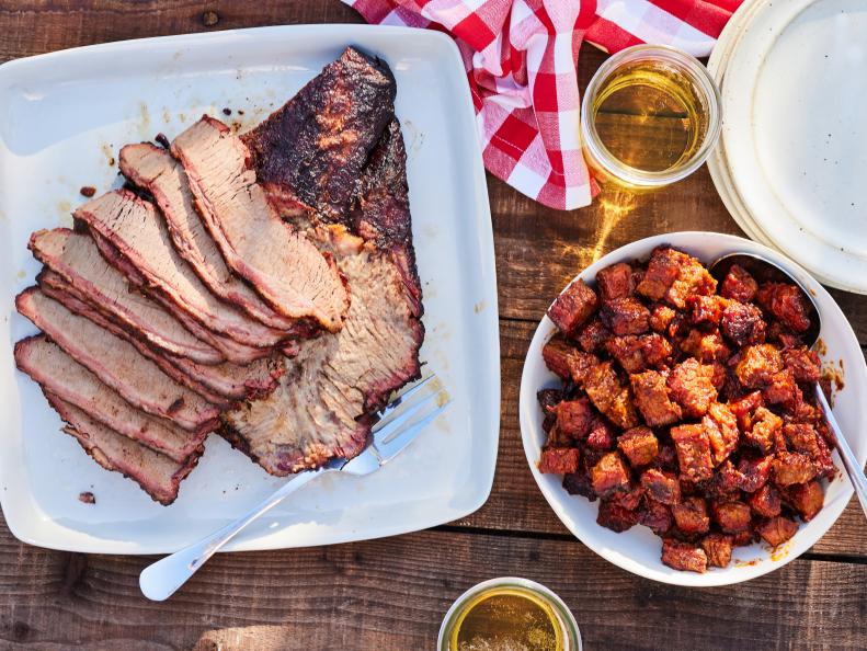 Sliced smoked brisket served on a white plate with a bowl of burnt ends, accompanied by glasses of beer and a checkered red napkin on a wooden table.